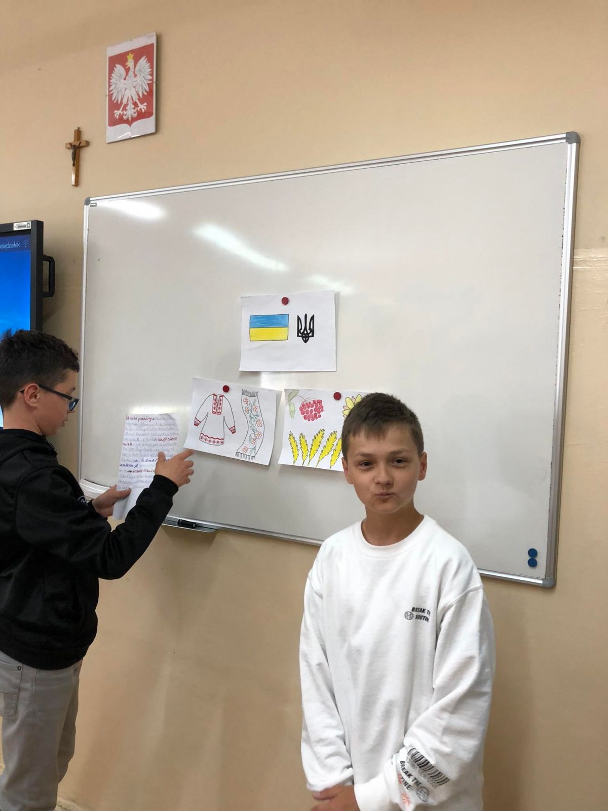 Na zdjęciach uczniowie z Ukrainy stoją przy tablicy na której wiszą samodzielnie wykonane plansze przedstawiające symbole Ukrainy.